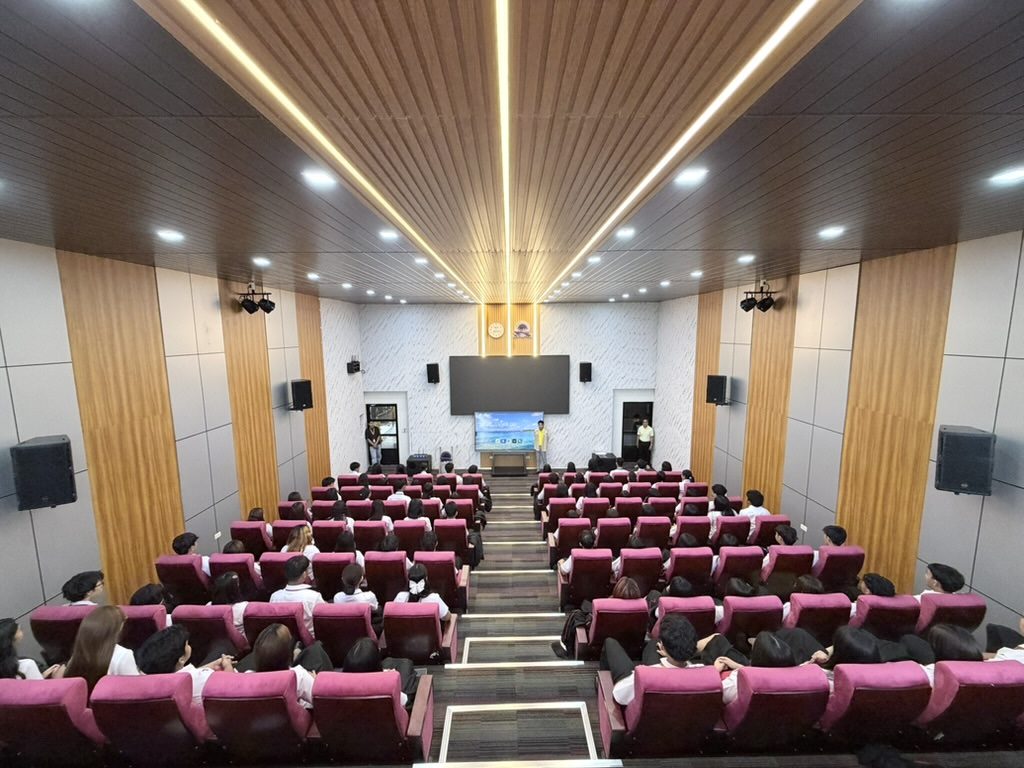 Empty Arcinas Hall photographed from the back, the centre aisle and gold ceiling lights both drawing toward the stage screen.